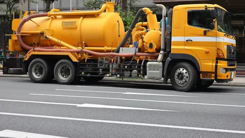 A yellow vacuum truck with a round tank, pump and suction hoses, parked on a city street